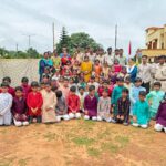 Large group of indian children in colorful traditional outfits posing with uniformed staff outdoors on a dusty school ground
